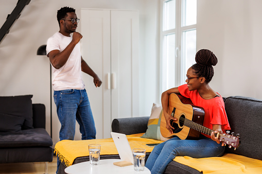 A cheerful woman is playing an acoustic guitar for her lovely boyfriend. A happy young African couple is sitting on the sofa in their apartment and enjoying the sounds of the guitar.