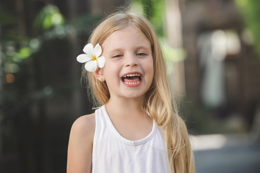 Candid outdoor portrait of adorable 5 years old girl with beautiful long blonde hair and frangipani flower behind ear, tropical vacation concept