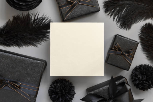 Blank card between Wrapped Gift Boxes, black paper flowers and feathers, mockup, copy space. Dark composition with square paper card for Birthday, Anniversary or black party present