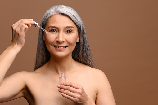 Happy beautiful middle-aged grey haired Asian woman using anti-aging serum, holding cosmetology product on brown background. Mature korean lady with hydrated skin holds pipette product near face