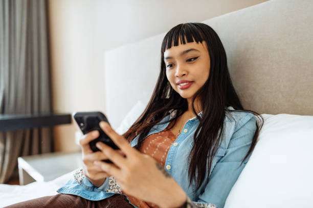 Mixed race Asian woman relaxing in bed and using smart phone
