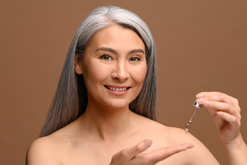 Charming Asian 40s lady using anti-aging complex with hyaluronic acid isolated on brown background. Sophisticated middle aged woman applies regenerating serum on the palm. Skincare concept