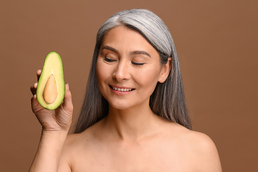 Portrait of beautiful middle-aged gray-haired Asian woman holding half of avocado isolated on brown background, 50s lady with naked shoulders standing with piece of fresh avocado and looking at it