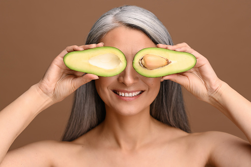 Beautiful Asian 50s mature grey-haired woman covering eyes with pieces of fresh avocado isolated on brown. Attractive mature lady recommends oil-rich foods. Healthy eating and lifestyle concept