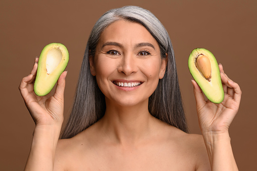 Cheerful joyful Asian 50s woman holding two half pieces of avocado, looks at camera and laughing. Middle-aged model with naked shoulders isolated on brown. Healthy lifestyle concept