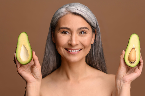 Portrait of middle-aged gray-haired half-naked Asian female holding halves of avocado in both hands, standing isolated on brown. Multiracial mature woman promoting healthy lifestyle and eating