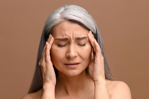 Depressed middle-aged Asian woman feels strong headache, upset female holding temples with hands isolated on brown, looking worried and anxious, suffering migraine, menopause