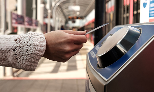 Female hand holding a card on contactless card reader machine in train station in London. There a blurry red train in the station. No cash public transport concept.