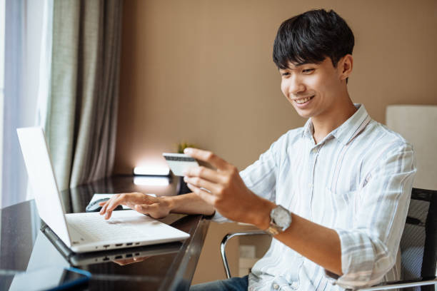 młody mężczyzna z azji południowo-wschodniej w pokoju hotelowym, korzystający z laptopa w celach biznesowych i telepracy - southeast asian ethnicity men laptop image type zdjęcia i obrazy z banku zdjęć