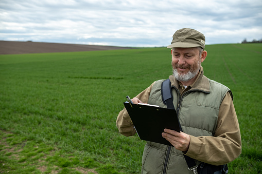 A cheerful farmer with a gray beard and gray hair in yellow-green work clothes and a laptop in a bag slung over his shoulder is writing on his clipboard in a green field.