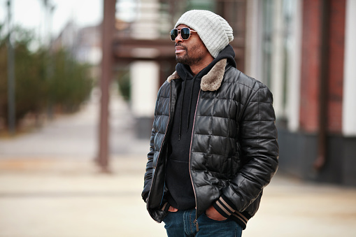 Portrait of young handsome black man, standing on cityscape, looking away, wearing leather jacket, knitted hat and sunglasses. Street style fashion