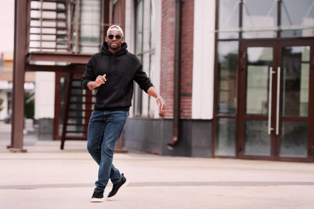 Cool Black man dancing on street of city alone, wears black hoodie, jeans and sneakers, baseball cap and sunglasses