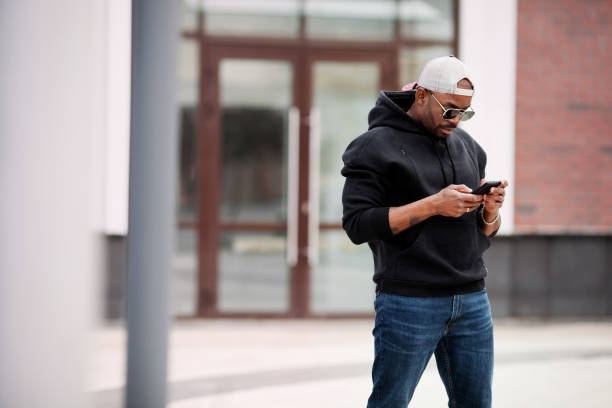 African American black man uses mobile phone, stands on city street, wears hoodie, baseball cap and sunglasses. Portrait Stylish male model looks at his phone