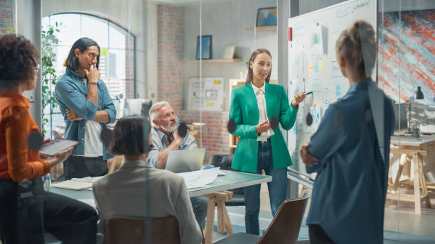 istock Female Project Manager Makes a Presentation for a Diverse Creative Team in Meeting Room in an Agency. Colleagues Gathering Around Conference Table and Discuss Business Development and Design. 1482198043