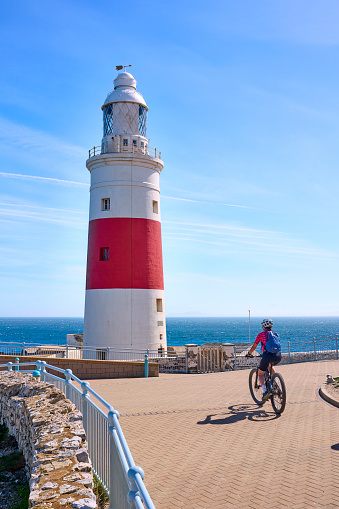 happy senior woman cycling with her electric mountain bike below the Lighthouse of Gibraltar, marking the Strait of Gibraltar, Andalusia, Spain and UK