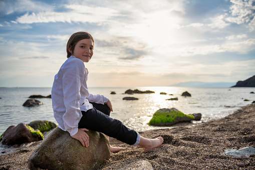 A serene seaside moment captured as a boy sits on a rocky shore, barefoot and happy, against a backdrop of a spectacular sunset and tranquil ocean