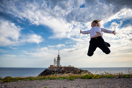 With a victorious spirit, a young boy jumps happily in his formal attire, against a scenic backdrop of a lighthouse and the ocean