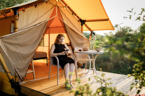 istock Happy Woman freelancer use phone and drinking cocktail call while relaxing on cozy glamping tent in sunny day. Luxury camping tent for outdoor summer holiday and vacation. Lifestyle concept 1482205728