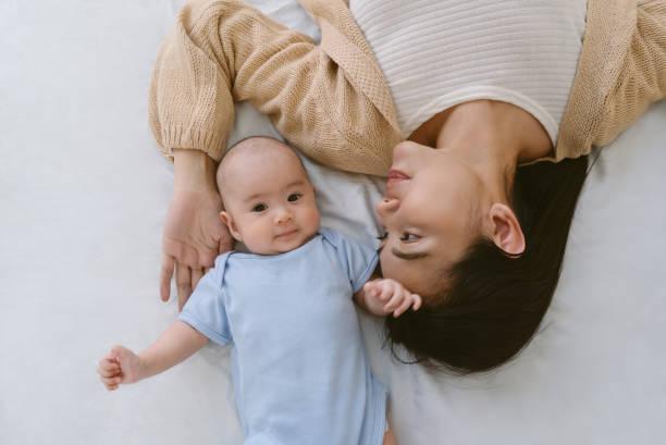 Beautiful young Asian mother and her adorable newborn baby lying on white bed at home. Newborn looking at camera and smiling. Mothercare is most important in baby life. Mother's day concept. hermosa joven madre asiática y su adorable bebé recién nacido acostado en una cama blanca en casa. recién nacido mirando a la cámara y sonriendo. el cuidado de la madre es más importante en la vida del bebé. concepto del día de la madre.