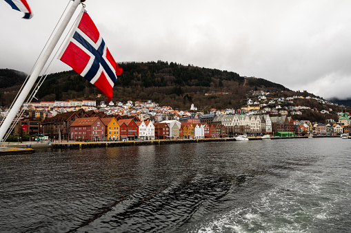Bergen, Norway - March 25, 2023: Bryggen Wharf in Bergen, Norway as viewed from across a boat on a cloudy day. Norwegian flag in the forefront and Mount Floyan in the background.