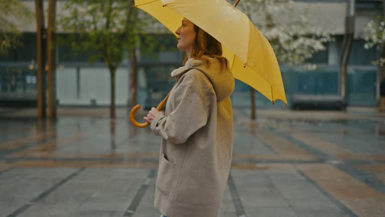 SLO MO Side view of Young woman walks with a yellow umbrella in the city on a rainy day