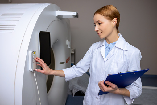 Female doctor wearing lab coat in magnetic resonance imaging or computed tomography room of modern clinic making diagnostics, using control panel.