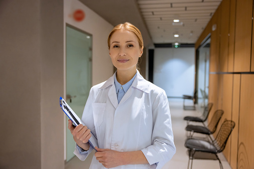 Attractive blonde young female doctor standing in hospital corridor and holding clipboard, working in nursing home.