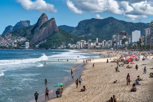 This photo captures the vibrant atmosphere of Ipanema Beach in Rio De Janeiro, Brazil. With its pristine white sand stretching along the azure waters of the Atlantic Ocean, this iconic beach is a magnet for tourists and locals alike. The photo showcases the beach's bustling boardwalk lined with palm trees, beach umbrellas, and colorful tents where visitors can enjoy snacks and drinks. In the distance, the towering mountains of Rio de Janeiro provide a stunning backdrop to the scene. This photo is perfect for travel brochures, websites, or any project that aims to capture the beauty and energy of one of the world's most famous beaches.