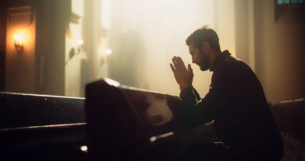 o jovem cristão senta-se piedosamente na majestosa igreja, com as mãos cruzadas depois de uma oração cruzada. ele busca orientação da fé e da espiritualidade. crente religioso no poder e no amor de deus - penance - fotografias e filmes do acervo