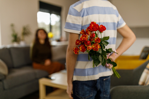 Rear view of caring teenage boy holding a beautiful bouquet of red roses behind his back, to surprise his loving mother while she is sitting on the sofa and working from home. Focus on foreground.