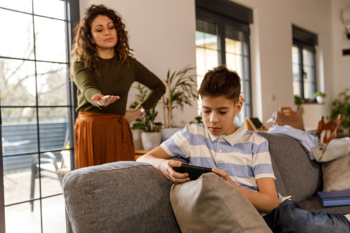 Selective focus shot of teenage boy lounging on the sofa, wasting time playing video games on smart phone. His angry mother standing behind him, hand on hip, looking at what he's doing and asking to hand her the phone.