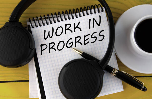 istock WORK IN PROGRESS - words on a white notepad on a yellow background with headphones, a cup of coffee and a pen 1482244202