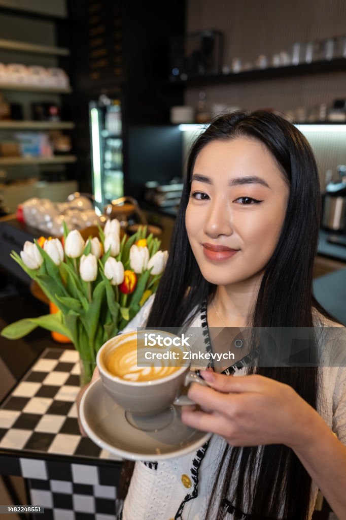 Smiling coffeehouse visitor with a caffeinated drink in her hand Waist-up portrait of a contented woman with a cup of cappuccino posing for the camera against the cafe interior 20-24 Years Stock Photo