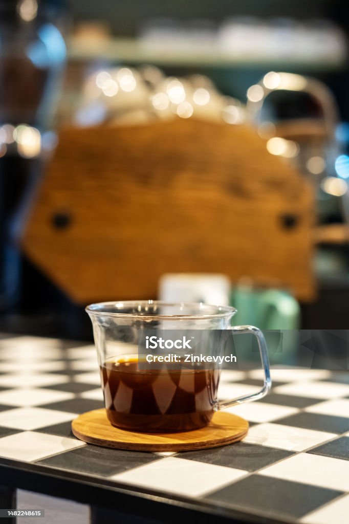 Caffeinated beverage served on the cafe table Close-up of a glass cup filled with black coffee placed on a handcrafted round wooden coaster in a coffeehouse Bar Counter Stock Photo