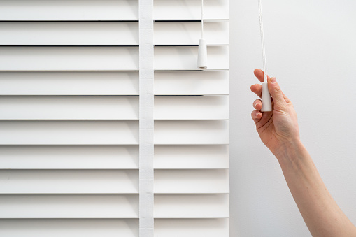 Woman hand pulls the adjustment string closing wooden jalousie slats in room. Cropped shot of female hand regulate light shade at louvers on plastic window