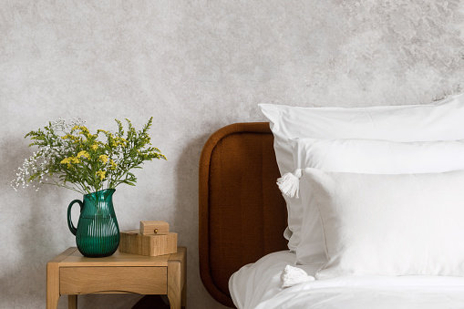 fresh bedding and cushions in cotton pillowcase on bed, glass vase with blossom flowers and storage jewelry box on wooden side table, detail of interior in hotel room