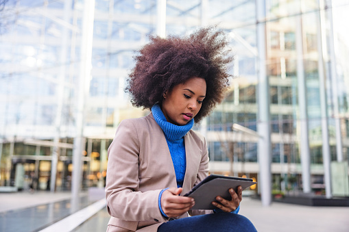 A busy black adult female entrepreneur using a tablet during her break outdoors. She is sitting outside an office building with big glass widows. The weather is cold thus she is wearing warm business casual clothes.