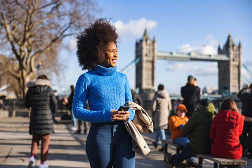 A black adult female tourist enjoying a sunny day outdoors and exploring London. She is walking by the river Thames near Tower bridge surrounded by other tourists. The weather is warm and sunny.