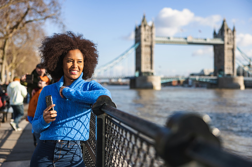 A happy black adult female waiting by the river Thames to meet a friend. She is leaning on the railing by the river and admiring the amazing view and the sunshine. In her hands, she is holding her phone to stay in contact with her late friend.