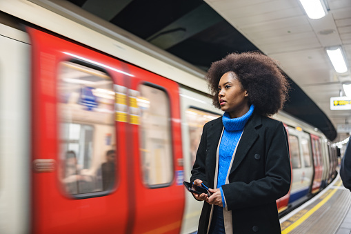A black adult female using London Underground to return home after a long day at work. She looks serious and tired. In her hands, there is a smartphone. In the background there is a train in motion blur.