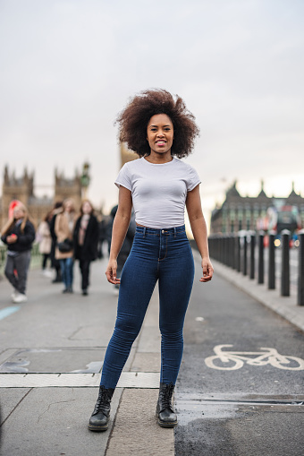 An attractive adult black female smiling at the camera while standing on a big bridge in London. She is wearing casual clothes. The weather is gloomy and typical for London.