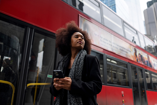 An adult black female standing on a sidewalk in London. She is looking around while holding a smartphone in her hands. In the background there is a typical red double-decker bus.