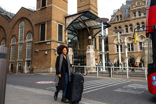 A happy adult black female tourist with a large suitcase walking from a railway station in London. She is on her way to the hotel. The woman is traveling alone and is excited to be able to explore the city. She is dressed in casual clothes.
