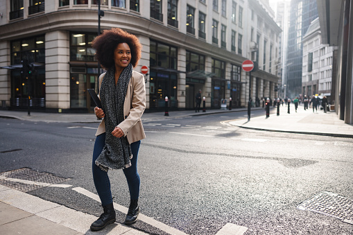 An adult black female walking on the streets of London alone and exploring the city. The is holding a tablet since she is returning from a business meeting on a sunny day. She is smiling and looks happy.