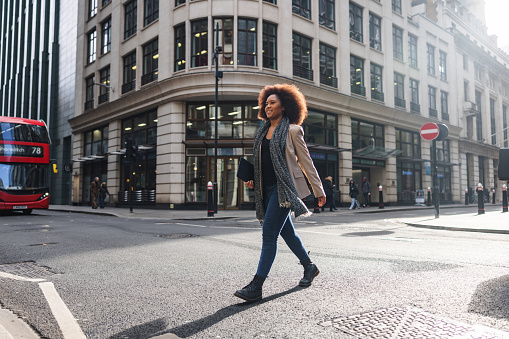 A happy adult black businesswoman crossing a street in London on her way to work. In the background, there is the typical red English double-decker. The woman is smiling and looks happy. She is carrying a tablet in her hands.
