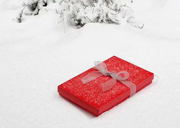A red gift box wrapped with a silver bow lies in the snow.