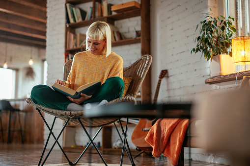 A young woman is at home relaxing and reading a book in her living room.