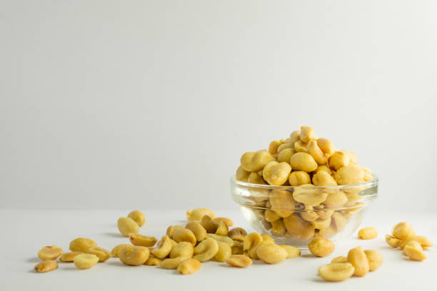 A still life photograph of salted and roasted peanuts in and around a small glass bowl isolated upon a white background