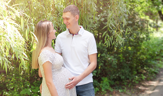 A young married couple is walking in the park. A man holds his hands on a pregnant woman's stomach.