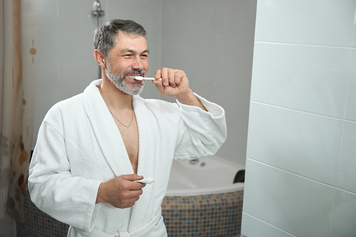 Adult man in white bathrobe standing in bathroom, looking in mirror and brushing teeth